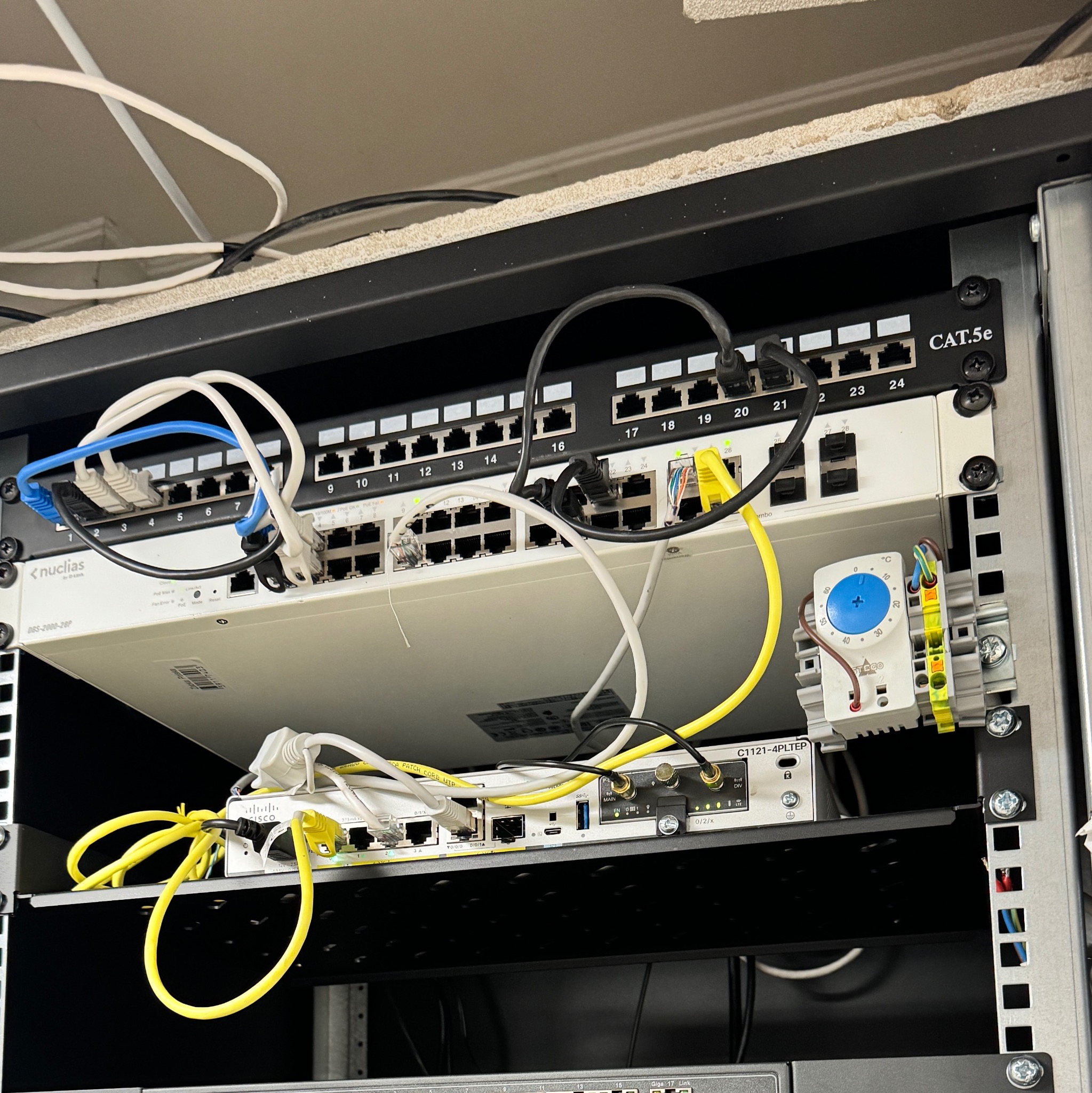 Szafa rack z zainstalowanymi urządzeniami sieciowymi: switche Nuclias i Cisco, patch panel Cat.5e oraz moduł Stego z niebieskim pokrętłem, okablowanie częściowo uporządkowane, widoczne kable...