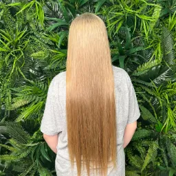 Długie, proste, blond włosy z delikatnym ombre na tle ściany z zielonych roślin.