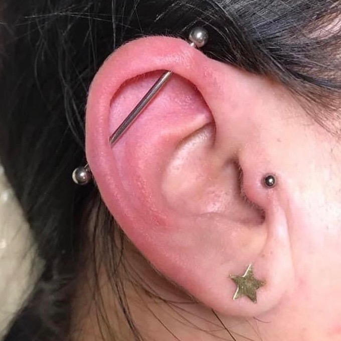 Ucho z trzema kolczykami: industrial barbell, tragus i złota gwiazdka w płatku ucha.