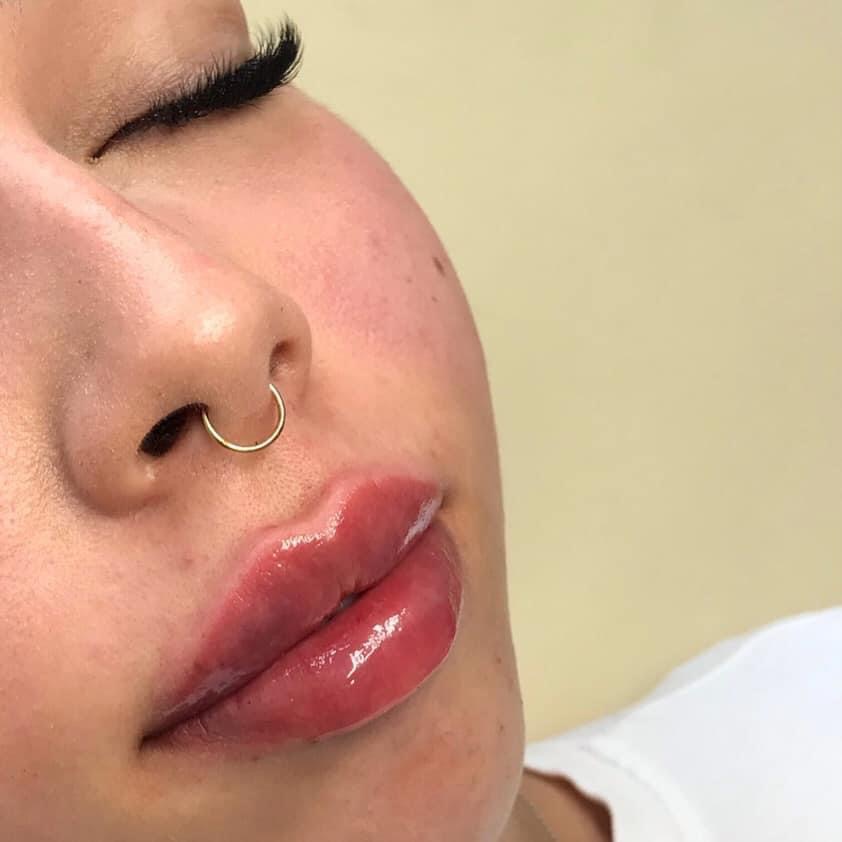 Zbliżenie na kobiecą twarz z błyszczącymi, powiększonymi ustami i złotym kolczykiem w przegrodzie nosowej, z widocznymi, przedłużonymi rzęsami.
