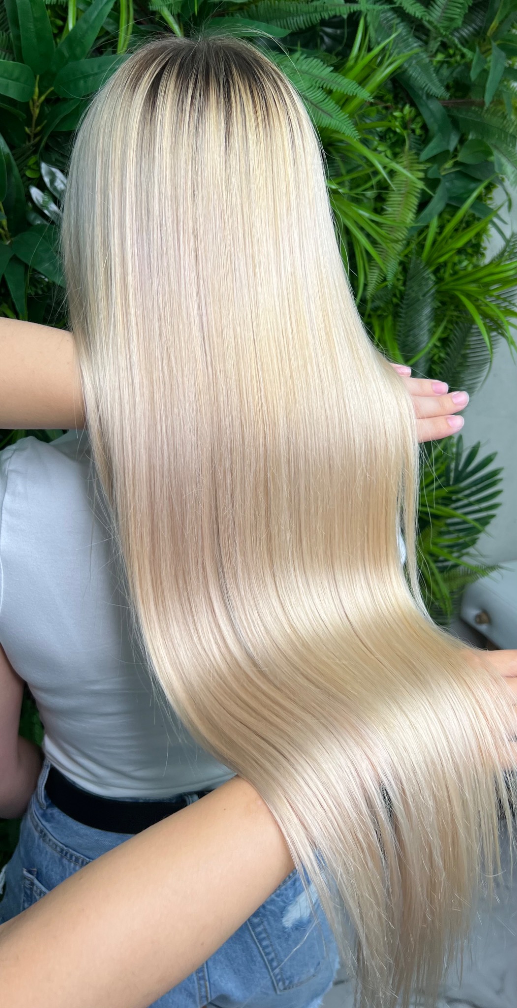 Długie, proste, platynowe blond włosy z ciemniejszym odrostem, prezentowane na tle ściany z zielonych roślin.