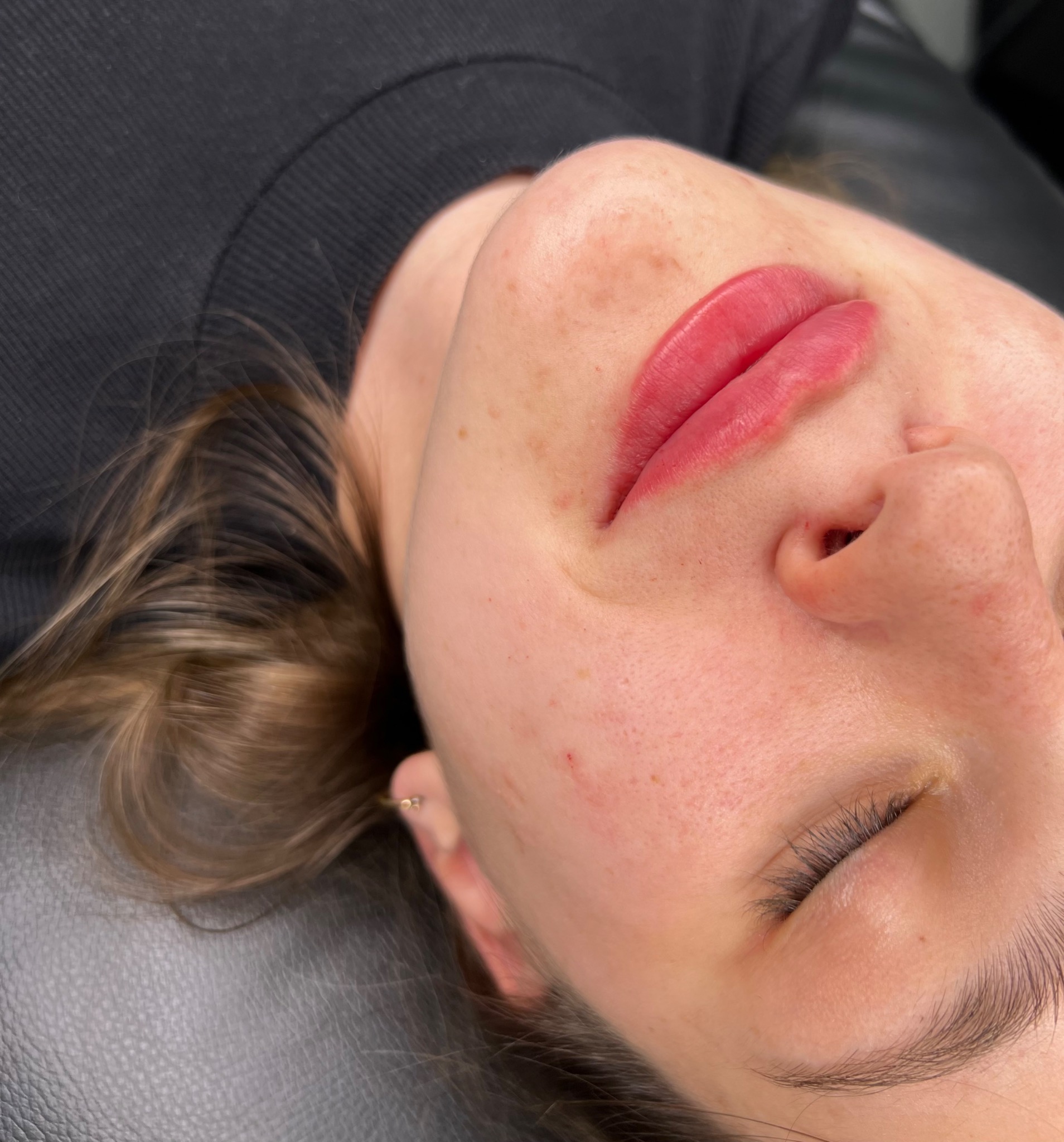 Zbliżenie na twarz kobiety leżącej na czarnym fotelu po zabiegu makijażu permanentnego ust, widoczne świeżo podkreślone, czerwone usta i zamknięte oko.