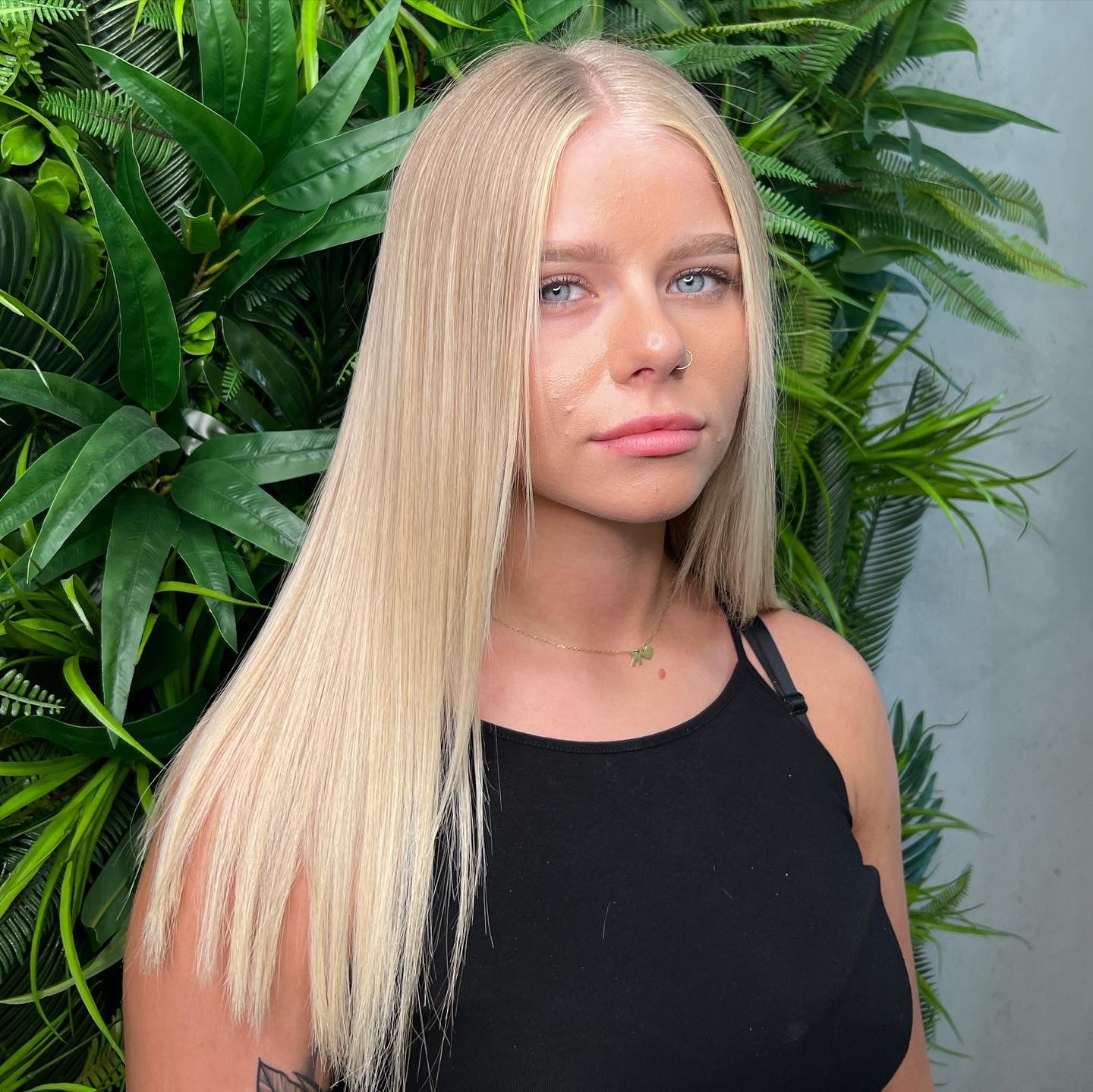 Portret młodej kobiety z długimi, prostymi, platynowymi włosami na tle ściany z zielonych roślin.