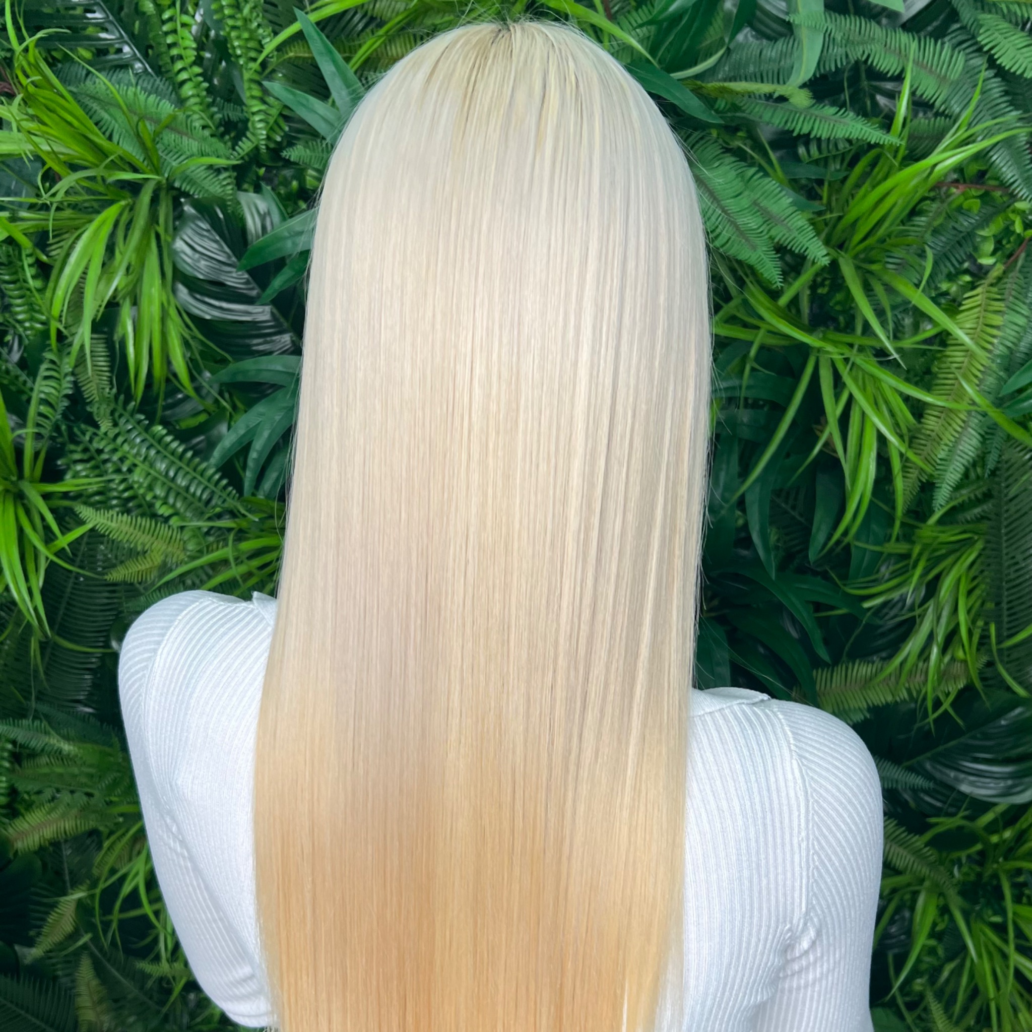 Długie, proste, platynowe włosy z delikatnym ombre w odcieniach blondu, na tle ściany z zielonych roślin.