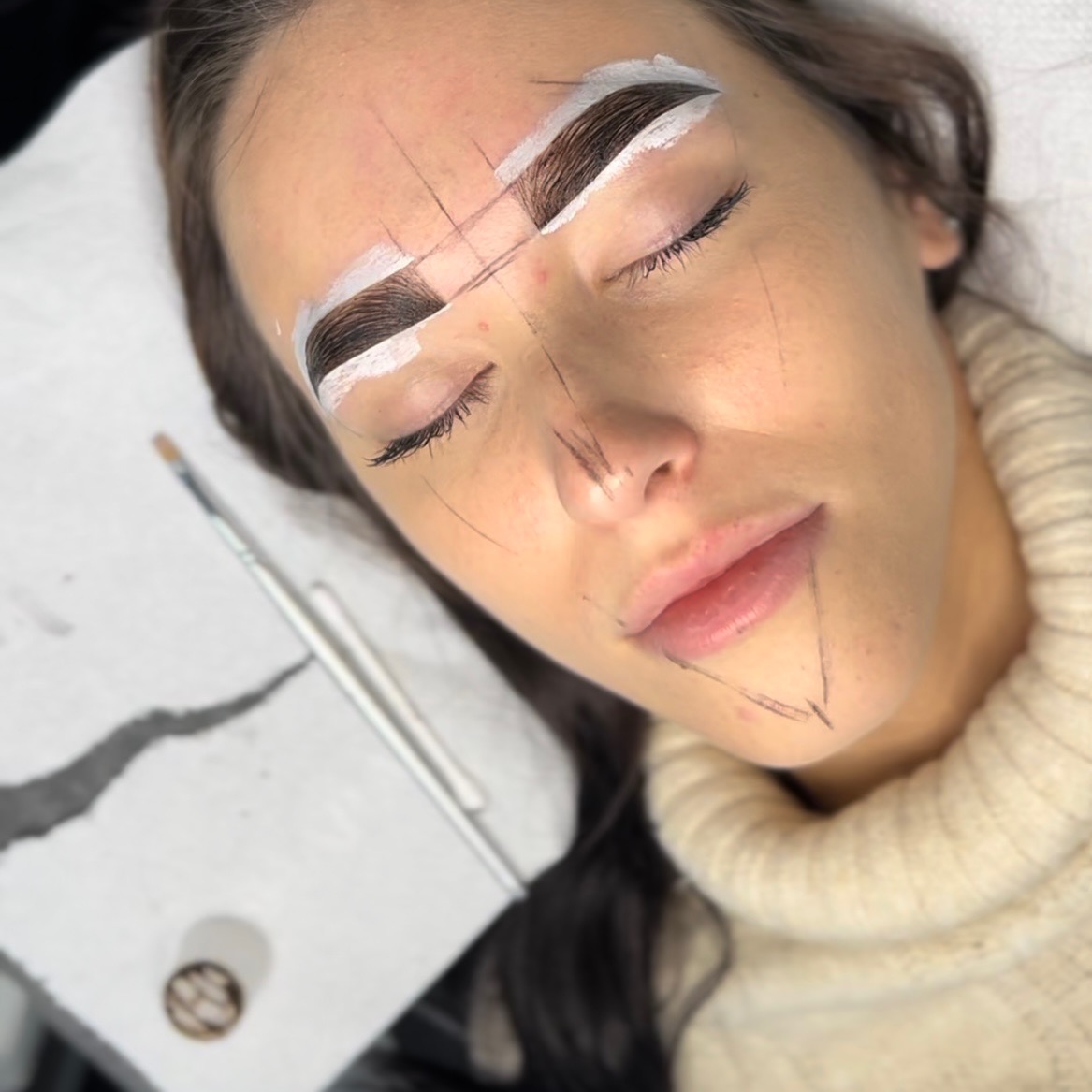 Kobieta z zamkniętymi oczami podczas zabiegu modelowania brwi z użyciem białej pasty i czarnych linii pomocniczych na twarzy, pędzelek i patyczki kosmetyczne obok.