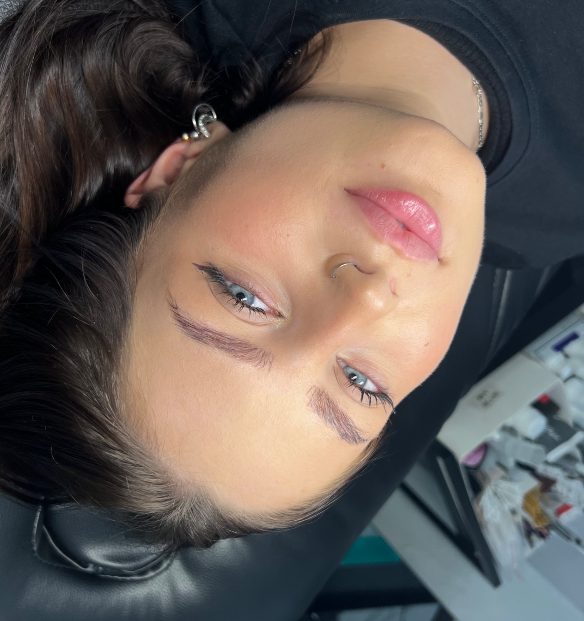 Twarz kobiety z delikatnym makijażem permanentnym brwi, widoczny kolczyk w nosie i uchu, leżąca na czarnym fotelu w salonie kosmetycznym.