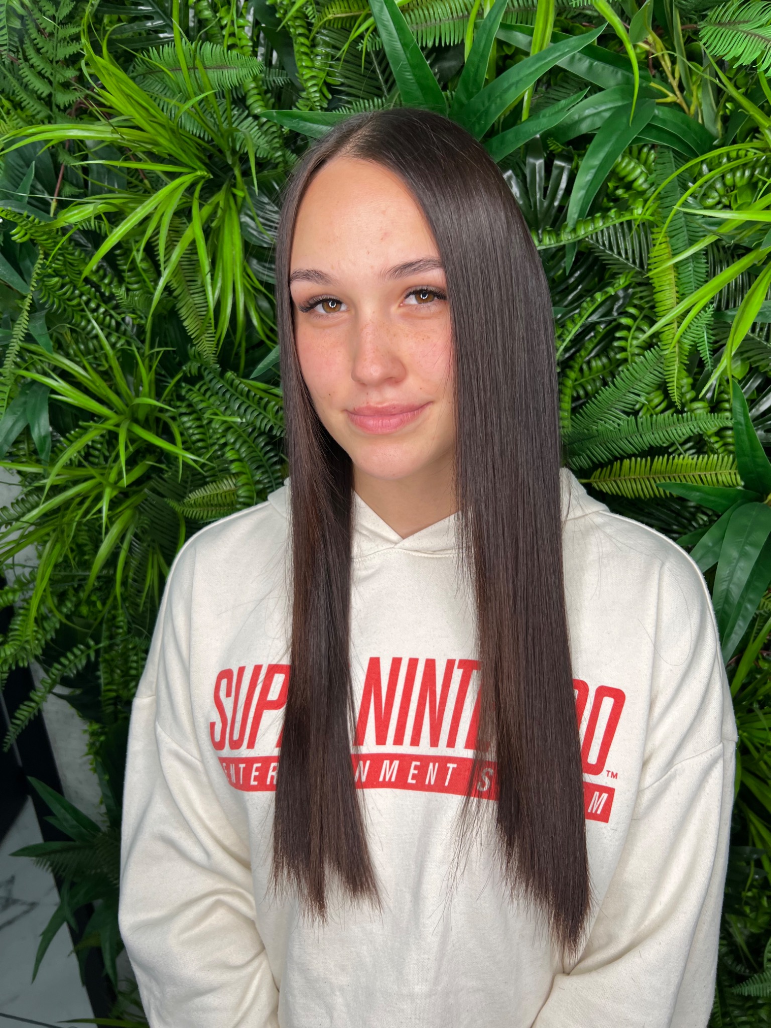 Portret kobiety z długimi, idealnie wyprostowanymi, brązowymi włosami na tle ściany z zielonych roślin.