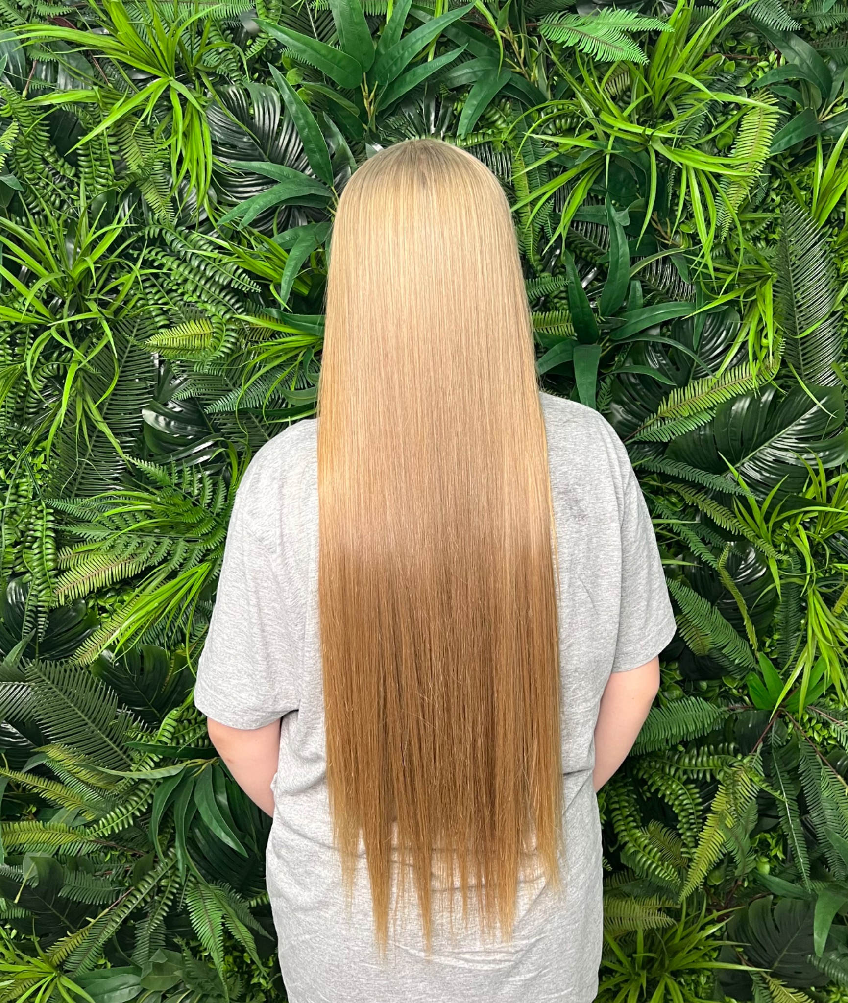 Długie, proste, blond włosy z delikatnym ombre na tle ściany z zielonych roślin.
