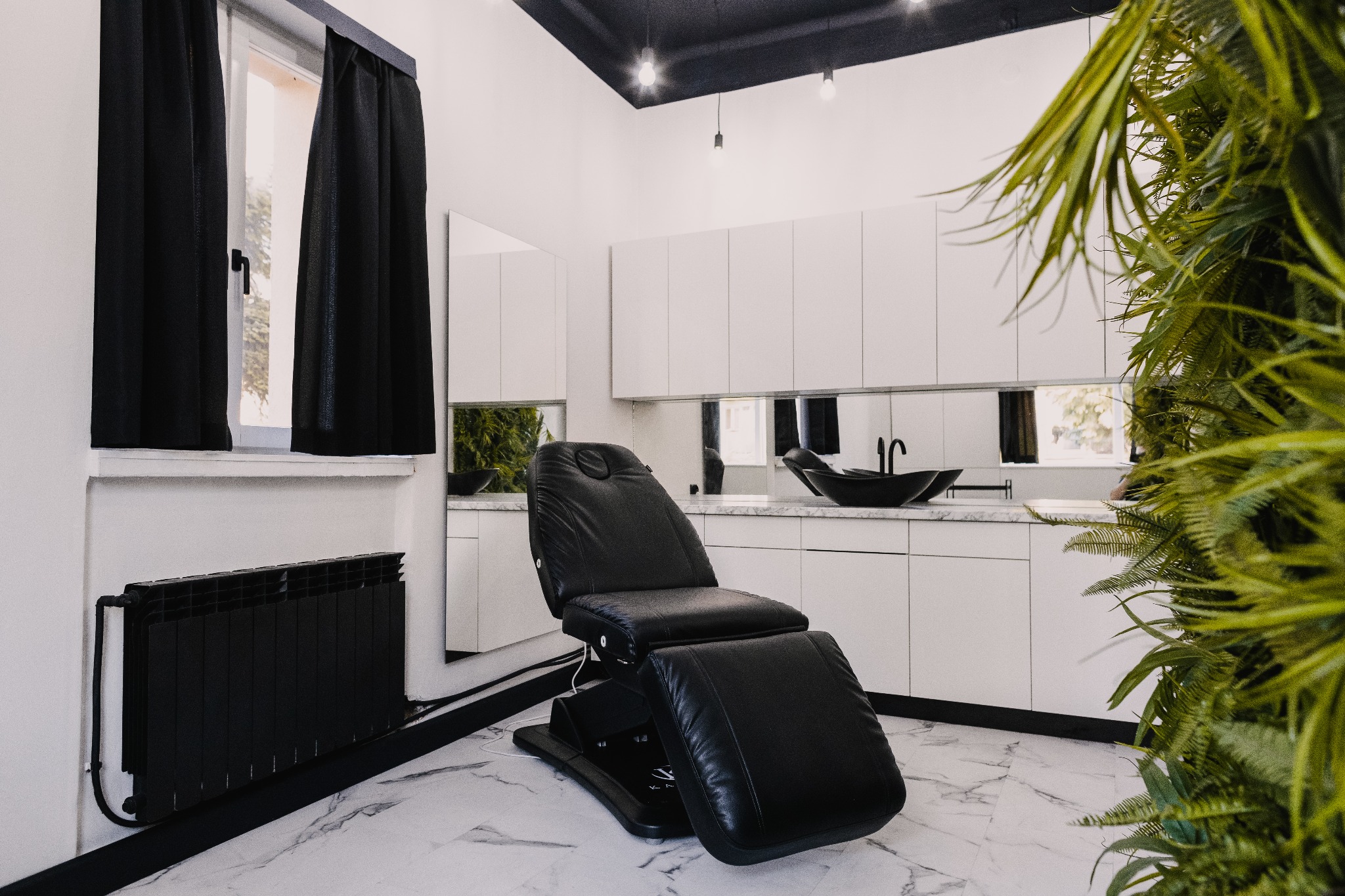 Nowoczesny gabinet kosmetyczny z czarnym fotelem zabiegowym, białymi szafkami, czarną umywalką i elementami zieleni w tle, oświetlony punktowymi lampami.