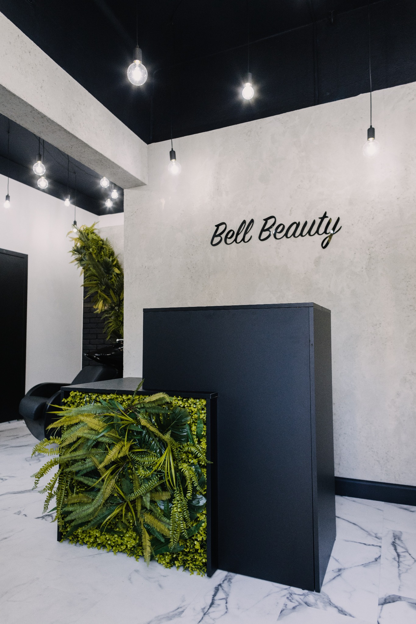 Nowoczesne wnętrze salonu z recepcją w kolorze czarnym, ozdobioną panelem z zielonych roślin, marmurowa podłoga i ściana z napisem 'Bell Beauty', oświetlenie punktowe.