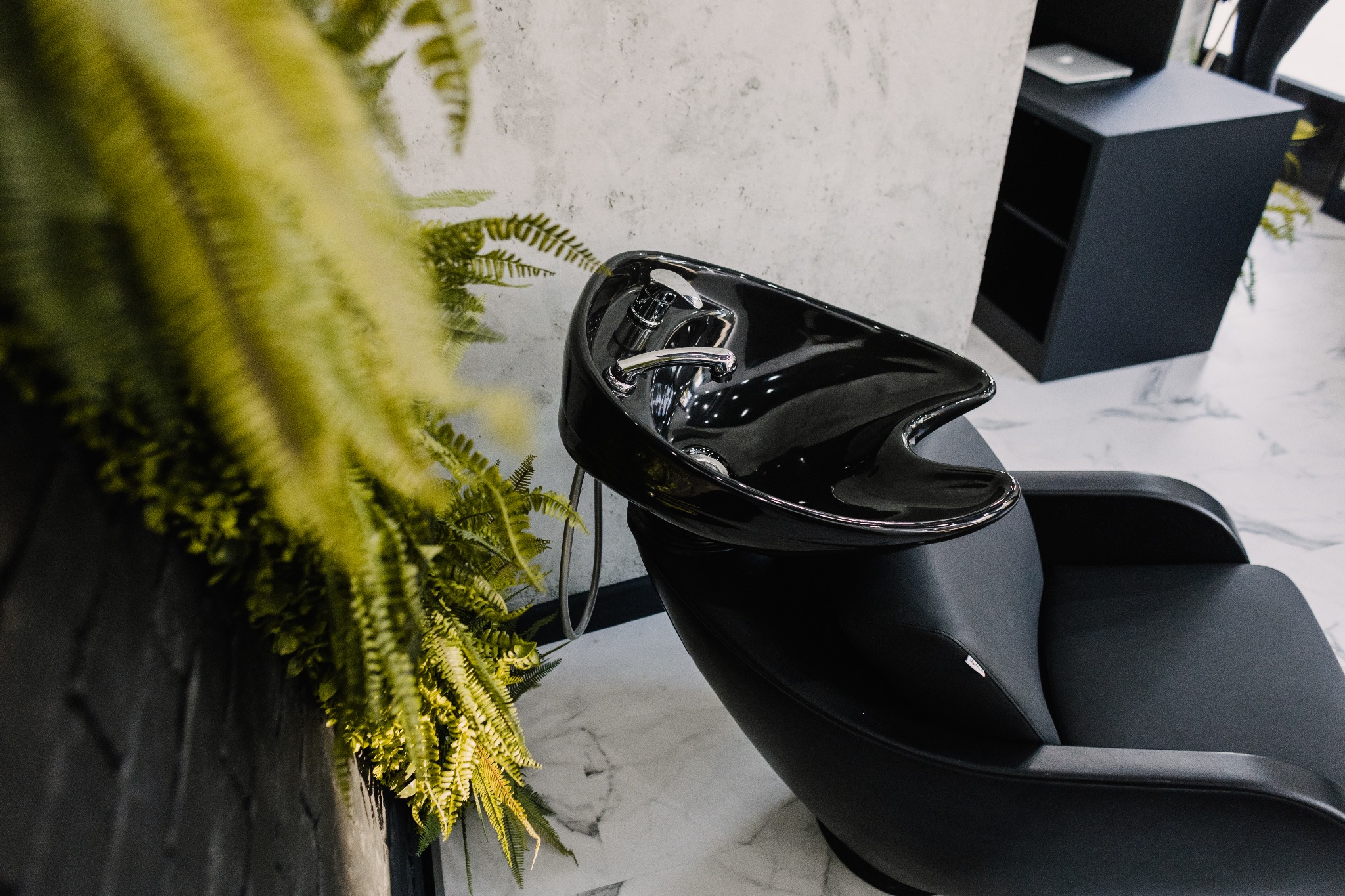 Czarne krzesło fryzjerskie z umywalką, widok z boku, z elementami dekoracyjnymi w postaci zielonych roślin, marmurowa podłoga, nowoczesny wystrój salonu.
