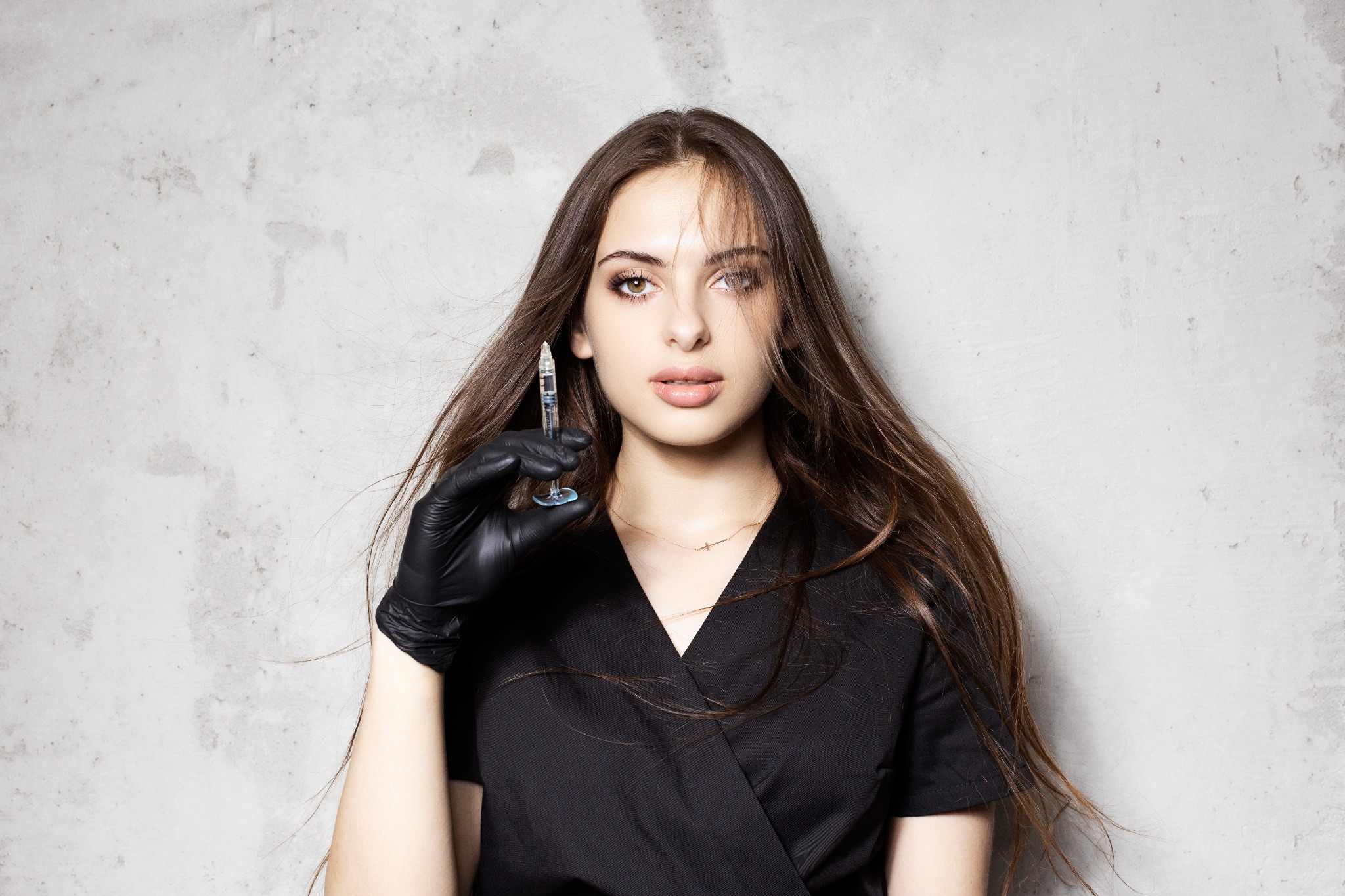 Kobieta z długimi brązowymi włosami, ubrana w czarny uniform i czarne rękawiczki, trzyma strzykawkę z przezroczystym płynem na tle szarej ściany.