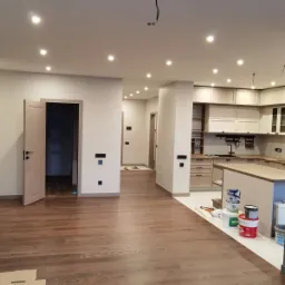 Wnętrze mieszkania w trakcie prac wykończeniowych, widoczna kuchnia z jasnymi meblami, drewniana podłoga, puszki z farbą i otwarte drzwi do innego pomieszczenia z czarnymi włącznikami na ścianie.