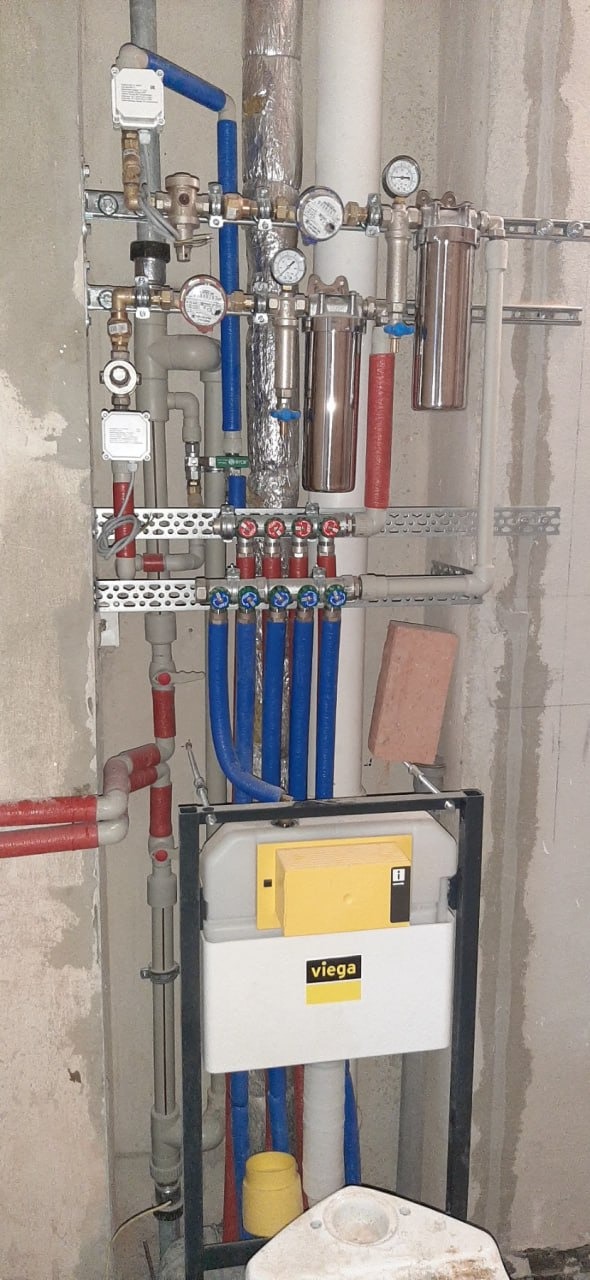 Instalacja wodno-kanalizacyjna w surowym stanie, widoczne rury miedziane, niebieskie i szare, filtry wody, manometry oraz stelaż podtynkowy WC marki Viega z żółtymi elementami, cegła podpierająca...