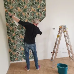 Mężczyzna w jeansach i czarnej koszuli przykłada arkusz tapety z motywem liści do ściany w jasnym pokoju, obok stoi drewniana drabina z gąbkami, na podłodze zielone wiadro i odkurzacz.