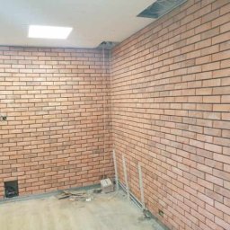 Usługi Remontowe Łukasz Kaszkowiak - Ściana wykończona cegiełkami klinkierowymi w jasnym odcieniu brązu z ciemną fugą, widoczne fragmenty instalacji elektrycznej i rur, surowy stan podłogi z pozostałościami po pracach wykończeniowych.