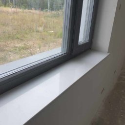 Usługi Remontowe Łukasz Kaszkowiak - Nowy, błyszczący biały parapet wewnętrzny pod szarym oknem z widokiem na pole, ściany przygotowane do malowania, widoczne ślady po szpachlowaniu.