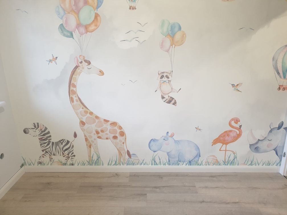 Pokój dziecięcy z ręcznie malowaną ścianą w motywy safari: żyrafa z balonami, zebra, szop pracz, hipopotam, flaming i nosorożec, utrzymane w pastelowej kolorystyce.