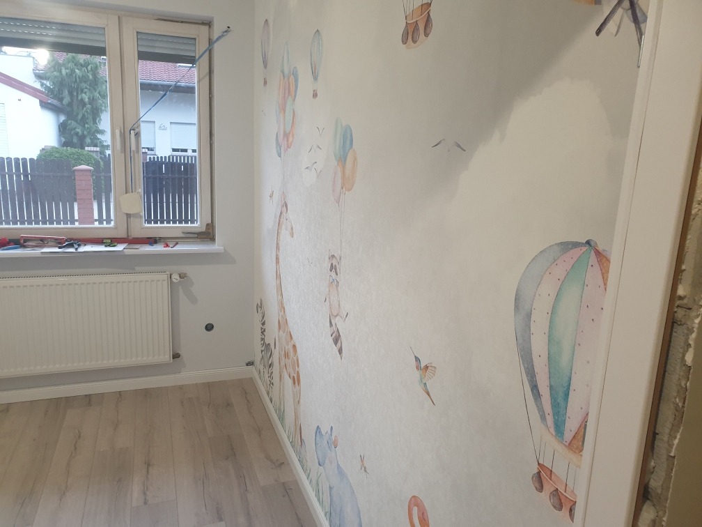 Pokój dziecięcy po dekoracji ścian tapetą z motywem zwierząt i balonów; widoczny fragment okna z narzędziami na parapecie.
