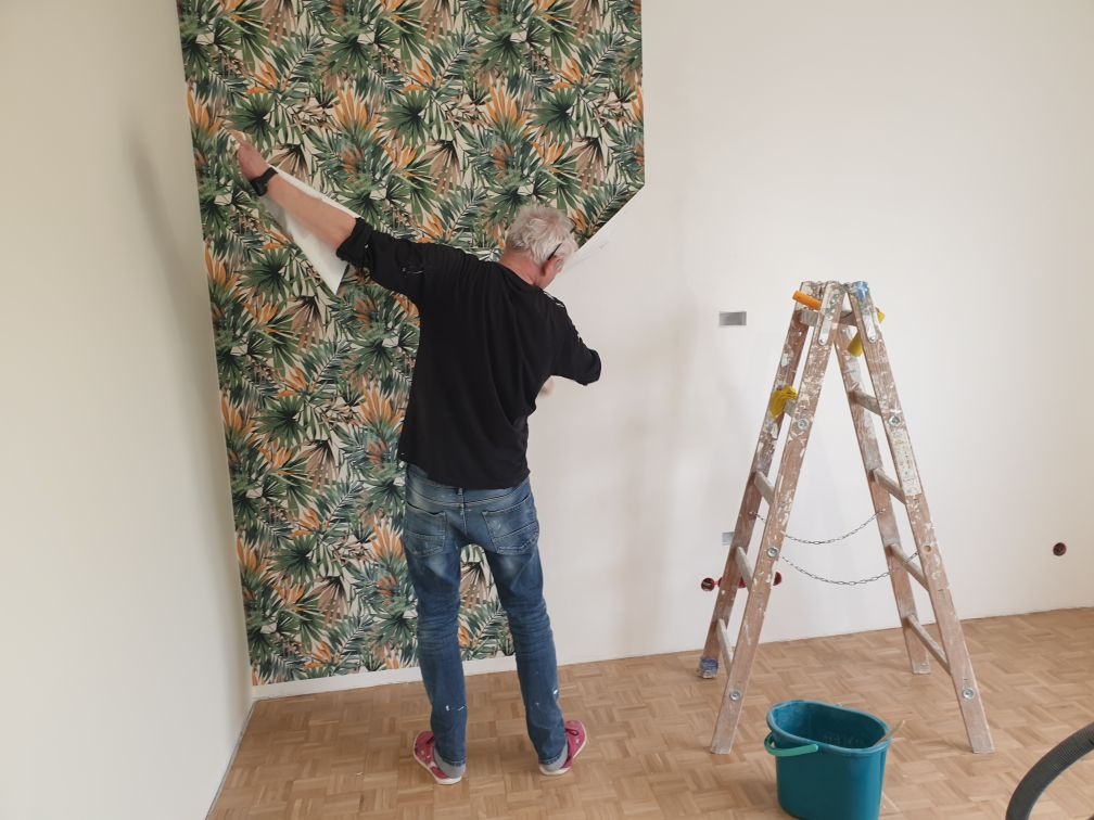 Mężczyzna w jeansach i czarnej koszuli przykłada arkusz tapety z motywem liści do ściany w jasnym pokoju, obok stoi drewniana drabina z gąbkami, na podłodze zielone wiadro i odkurzacz.