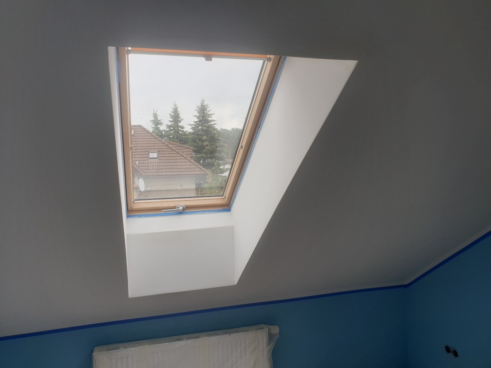 Poddasze z oknem dachowym po malowaniu, zabezpieczone taśmą malarską, widok na dom z brązowym dachem i drzewa za oknem.