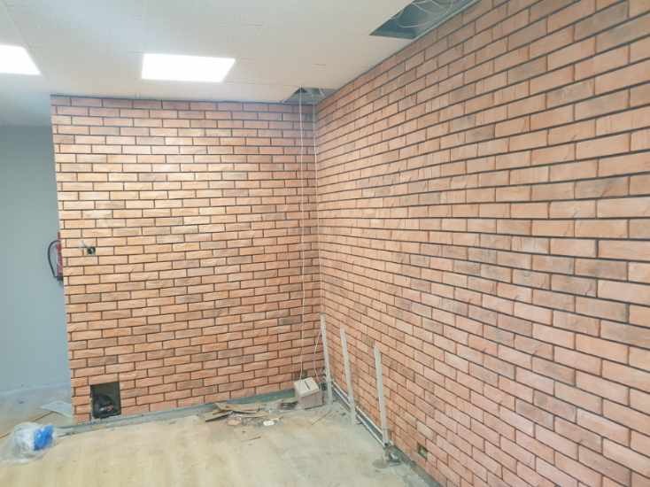 Ściana wykończona cegiełkami klinkierowymi w jasnym odcieniu brązu z ciemną fugą, widoczne fragmenty instalacji elektrycznej i rur, surowy stan podłogi z pozostałościami po pracach wykończeniowych.