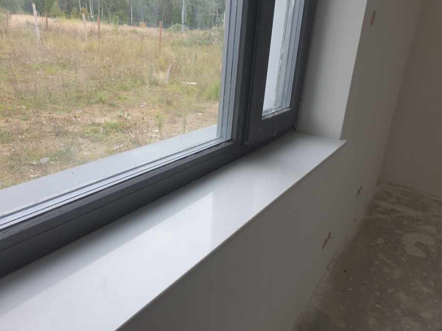 Nowy, błyszczący biały parapet wewnętrzny pod szarym oknem z widokiem na pole, ściany przygotowane do malowania, widoczne ślady po szpachlowaniu.
