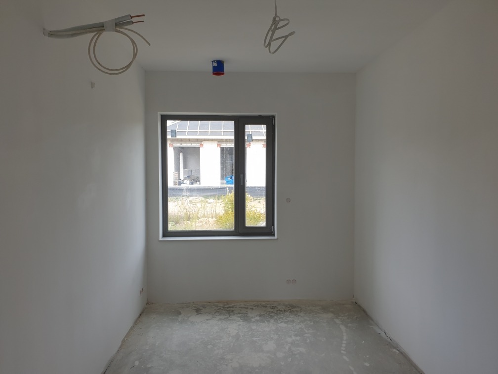 Pusty pokój z oknem i wyprowadzonymi przewodami elektrycznymi na suficie i ścianie, podłoga w stanie surowym.