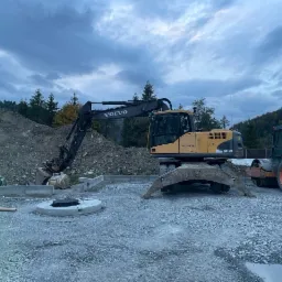 Żółta koparka Volvo EW160C na placu budowy z walcem drogowym ABG w tle, widoczny wykop i betonowe obrzeża, pochmurne niebo.