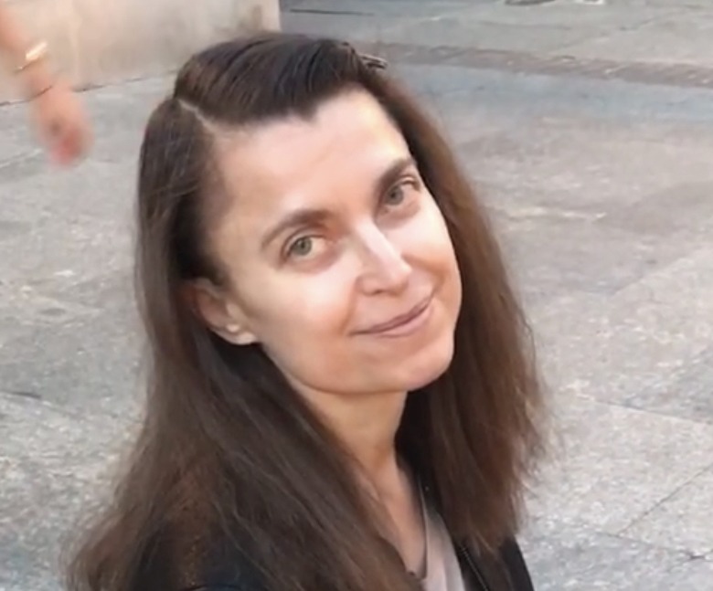 Portret kobiety z długimi brązowymi włosami, uśmiechającej się do kamery na tle brukowanej powierzchni, z dłonią w lewym górnym rogu kadru.