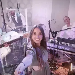 Zespół muzyczny podczas występu, widoczna wokalistka robiąca selfie z zespołem, perkusista za przezroczystą osłoną, klawiszowiec i basista.
