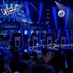 Scena programu The Voice of Poland z widownią, jurorami w obrotowych fotelach i wokalistką na podeście, oświetlona niebieskimi reflektorami.