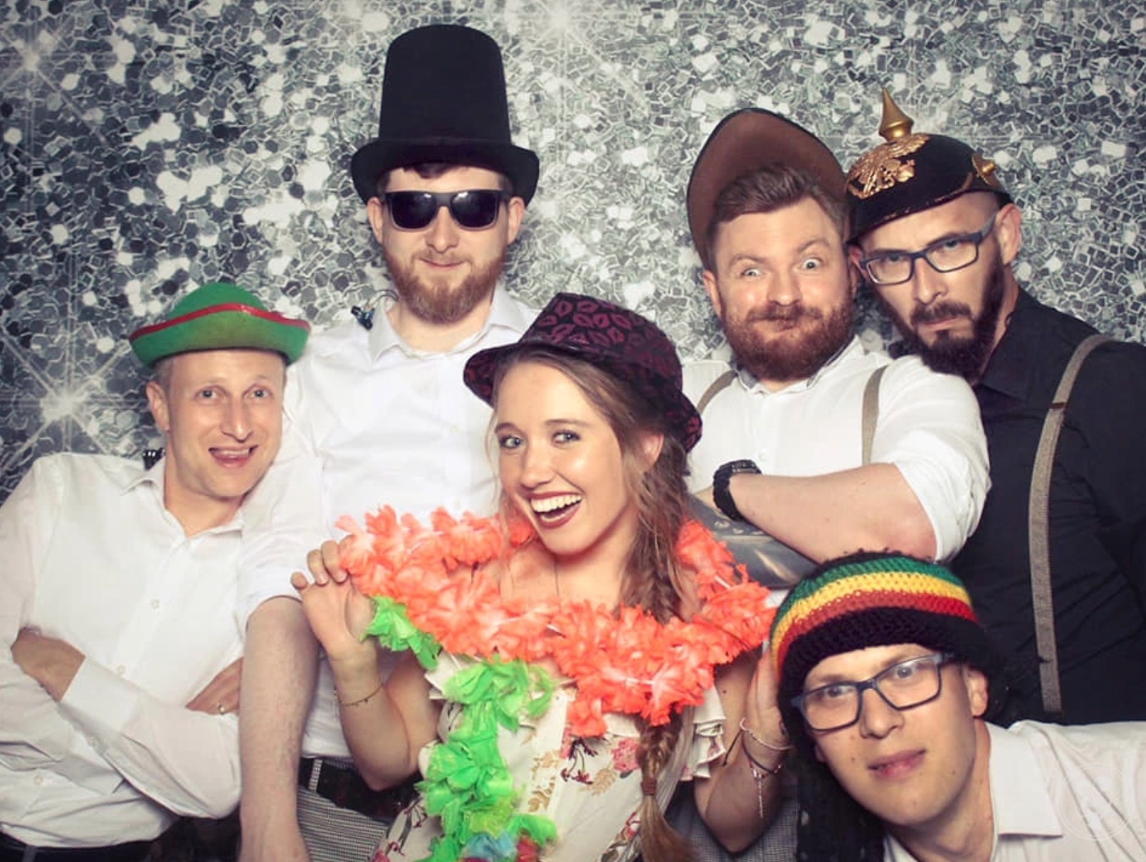 Grupa sześciu osób w przebraniach pozuje do zdjęcia na tle srebrnej, błyszczącej ściany. Widoczny cylinder, hawajski naszyjnik, czapka rasta i hełm z pikiem.