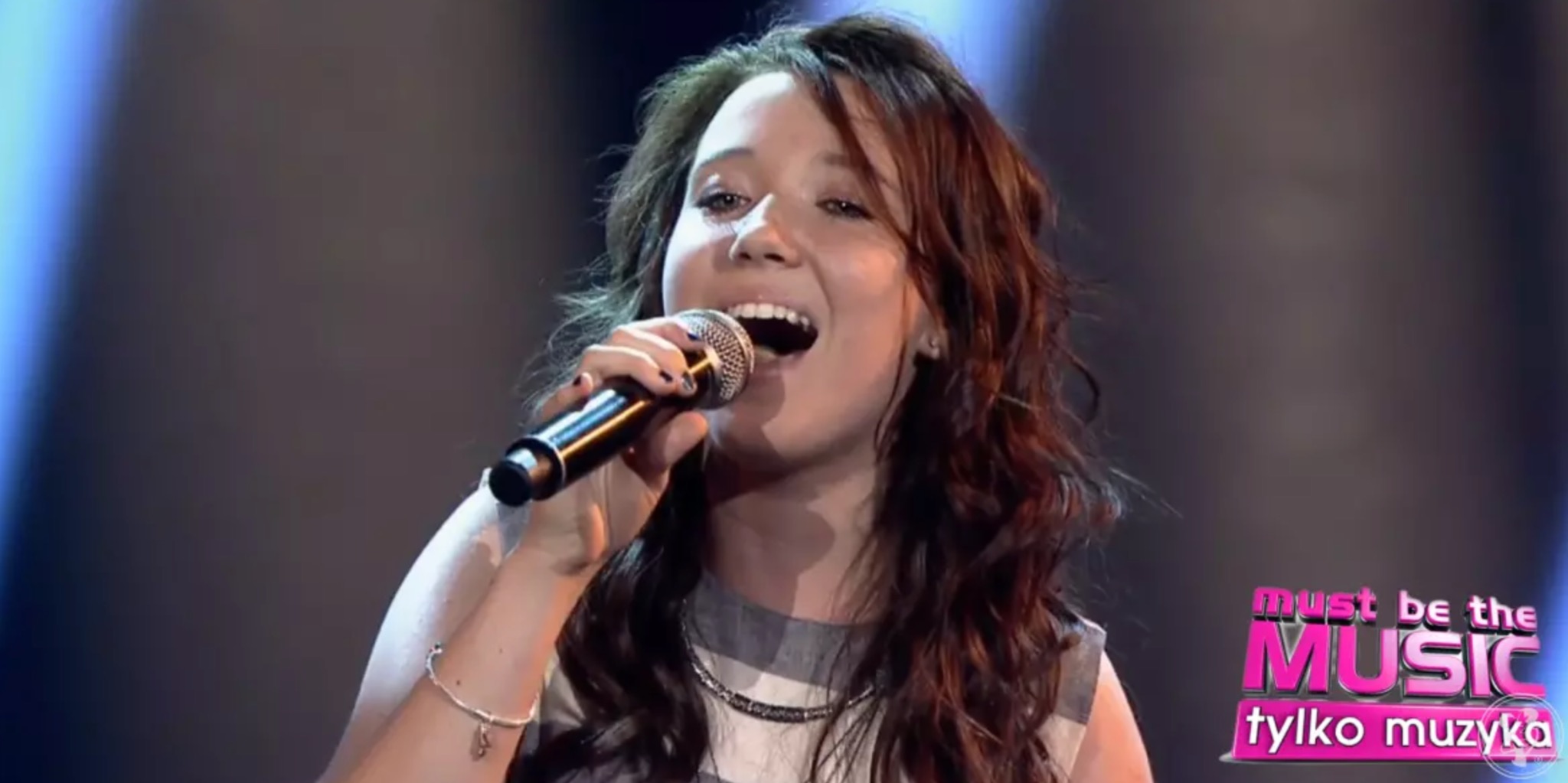 Wokalistka z Wodzisławia Śląskiego śpiewa do mikrofonu na scenie, w tle logo programu 'Must be the Music'.