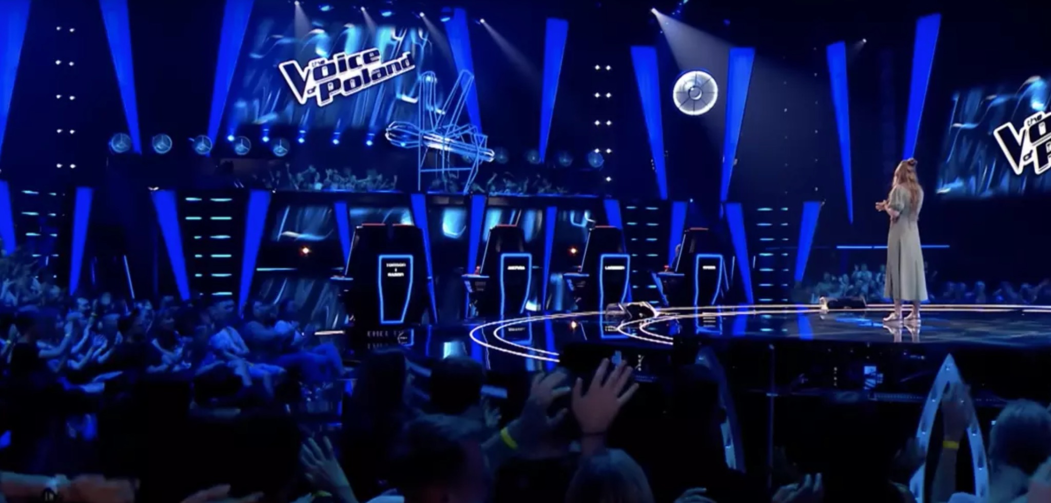 Scena programu The Voice of Poland z widownią, jurorami w obrotowych fotelach i wokalistką na podeście, oświetlona niebieskimi reflektorami.