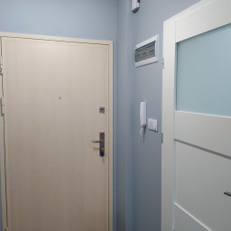 JW - Przedpokój po odświeżeniu: jasna farba na ścianach, beżowe drzwi wejściowe z wieszakiem na ubrania, białe drzwi wewnętrzne z przeszkleniem, skrzynka elektryczna na ścianie.