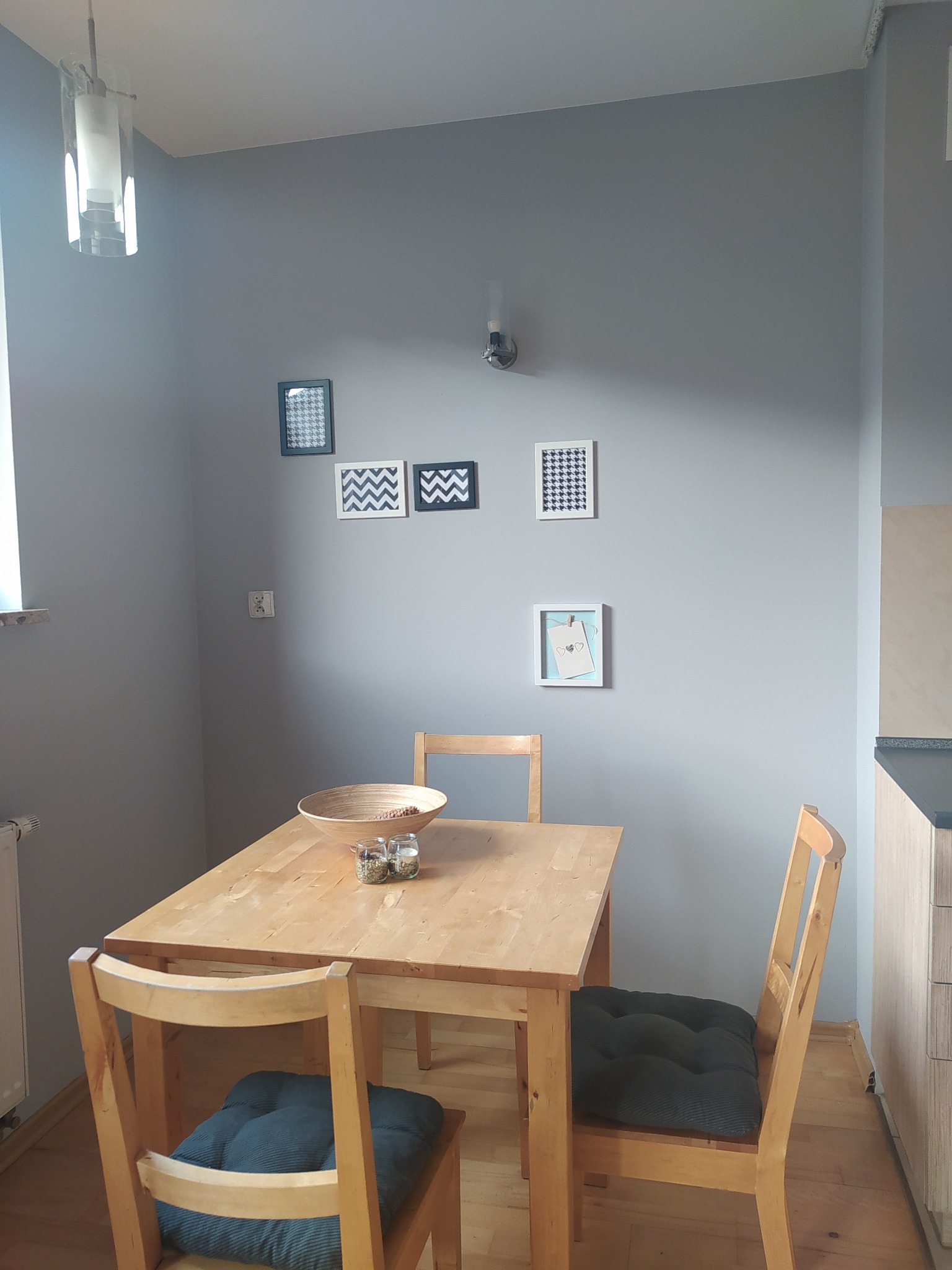 Szare wnętrze jadalni z drewnianym stołem, krzesłami z poduszkami, dekoracjami ściennymi i nowoczesną lampą wiszącą.