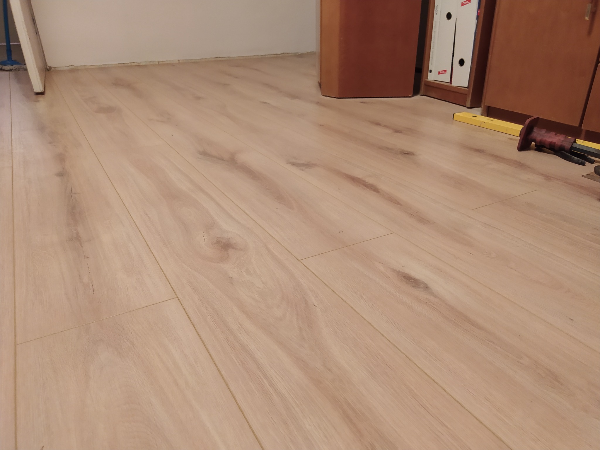 Jasna, drewnopodobna podłoga z paneli, widoczne narzędzia (poziomica, dłuto) przy krawędzi mebla, po prawej stronie kadru.