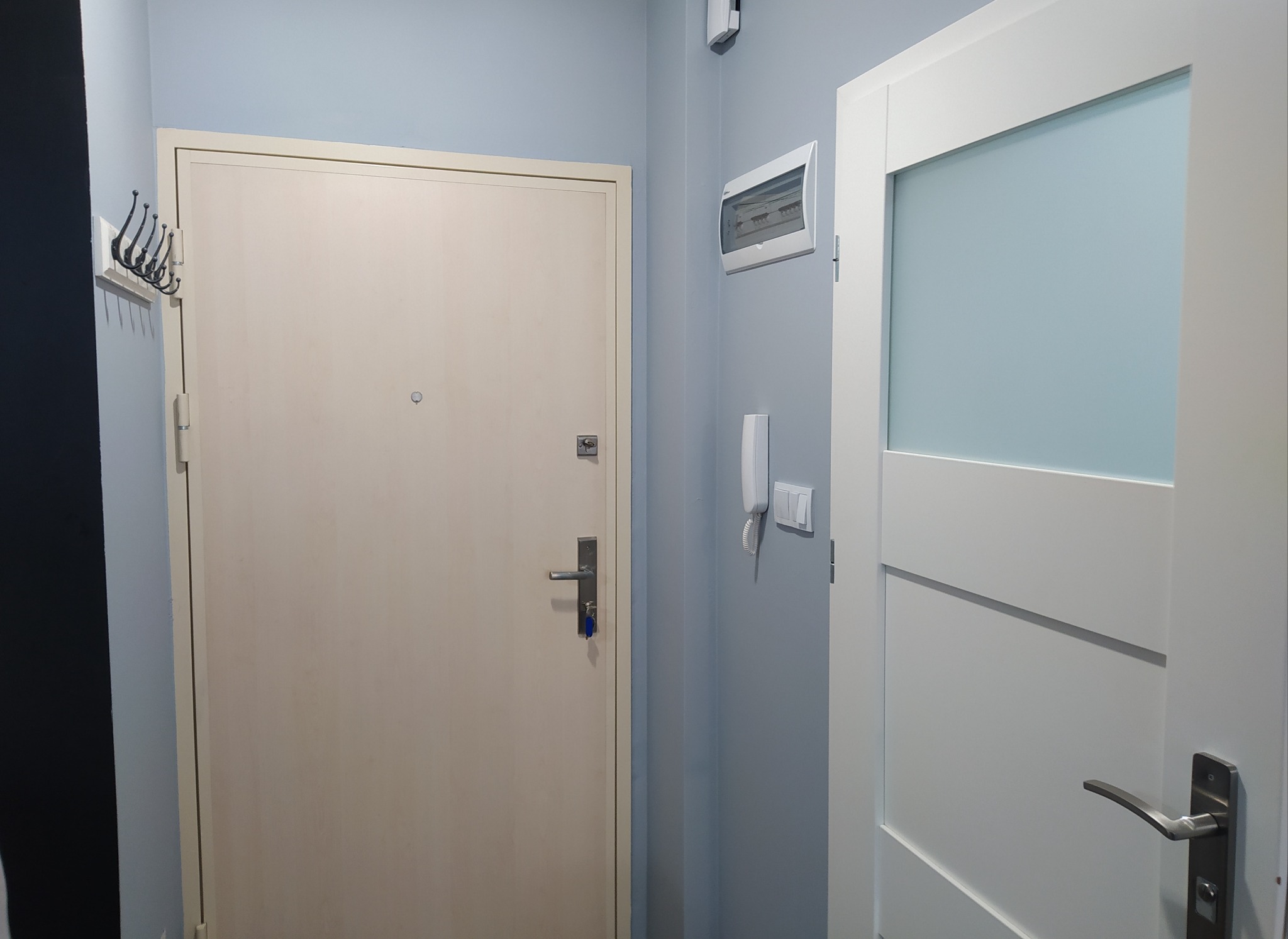 Przedpokój po odświeżeniu: jasna farba na ścianach, beżowe drzwi wejściowe z wieszakiem na ubrania, białe drzwi wewnętrzne z przeszkleniem, skrzynka elektryczna na ścianie.