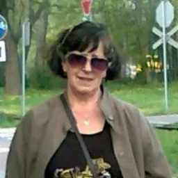 Portret kobiety w okularach przeciwsłonecznych, stojącej na tle znaków drogowych i drzew. Kobieta ma ciemne włosy i ubrana jest w brązową kurtkę.