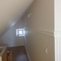 Poddasze po remoncie: białe ściany z boazerią i listwami, drewniana podłoga, okno w skosie dachu, oświetlenie punktowe.