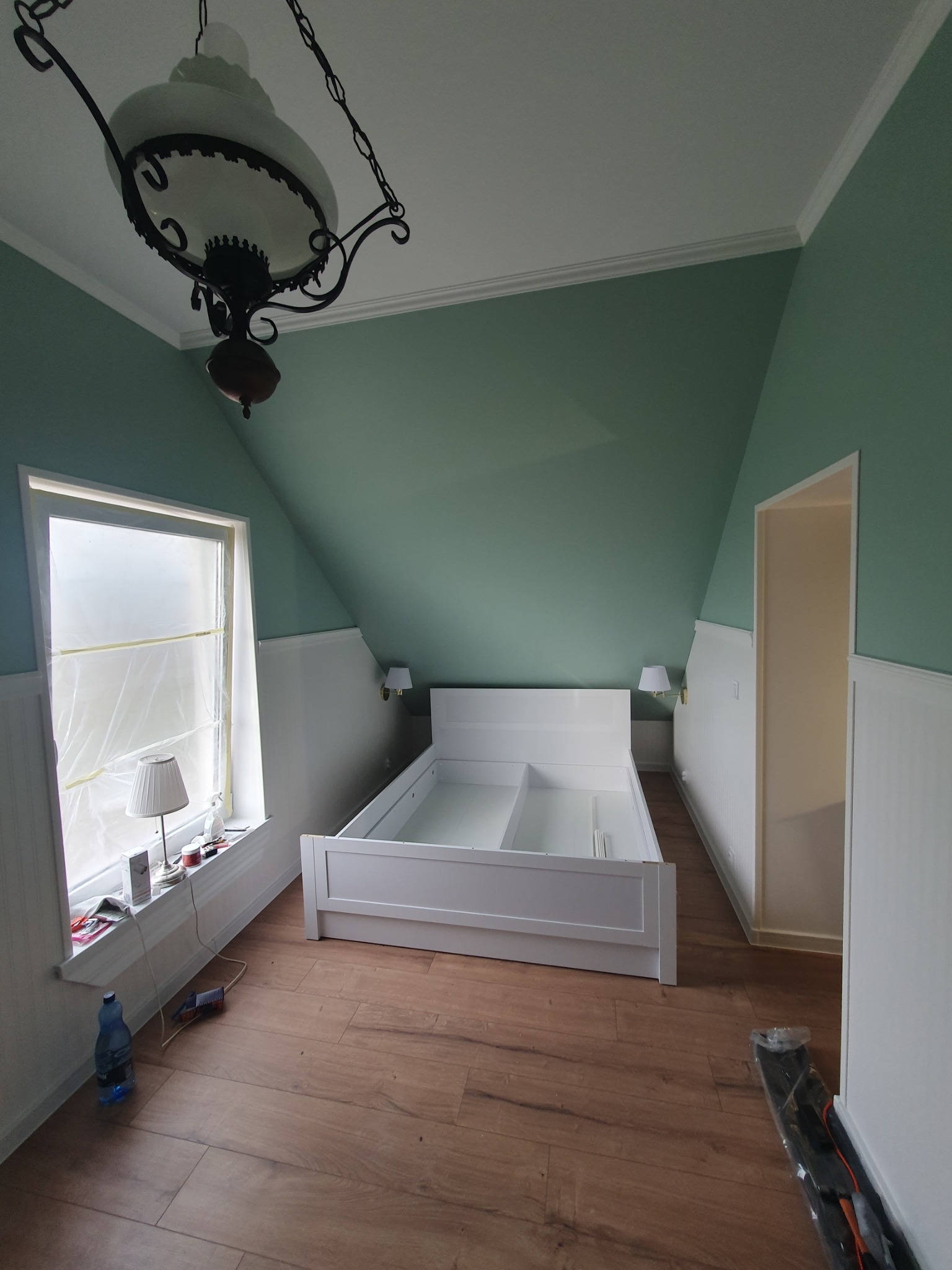 Pokój na poddaszu w trakcie wykańczania: białe panele ścienne, zielona górna część ścian, drewnopodobne panele podłogowe, biała rama łóżka bez materaca, okno zabezpieczone folią malarską, wisząca...