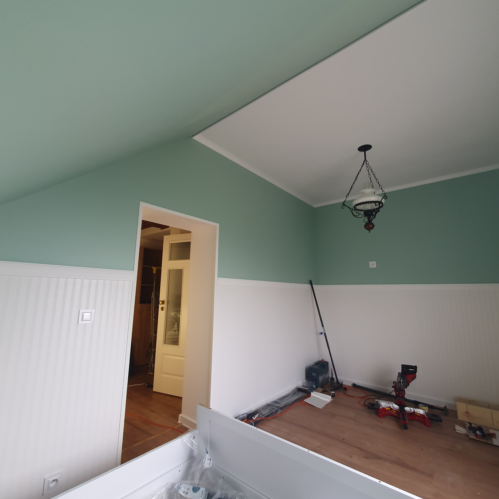 Pokój w trakcie malowania ścian na zielono i montażu białej boazerii, widoczna lampa w stylu retro, narzędzia do cięcia paneli podłogowych oraz otwarte drzwi do innego pomieszczenia.