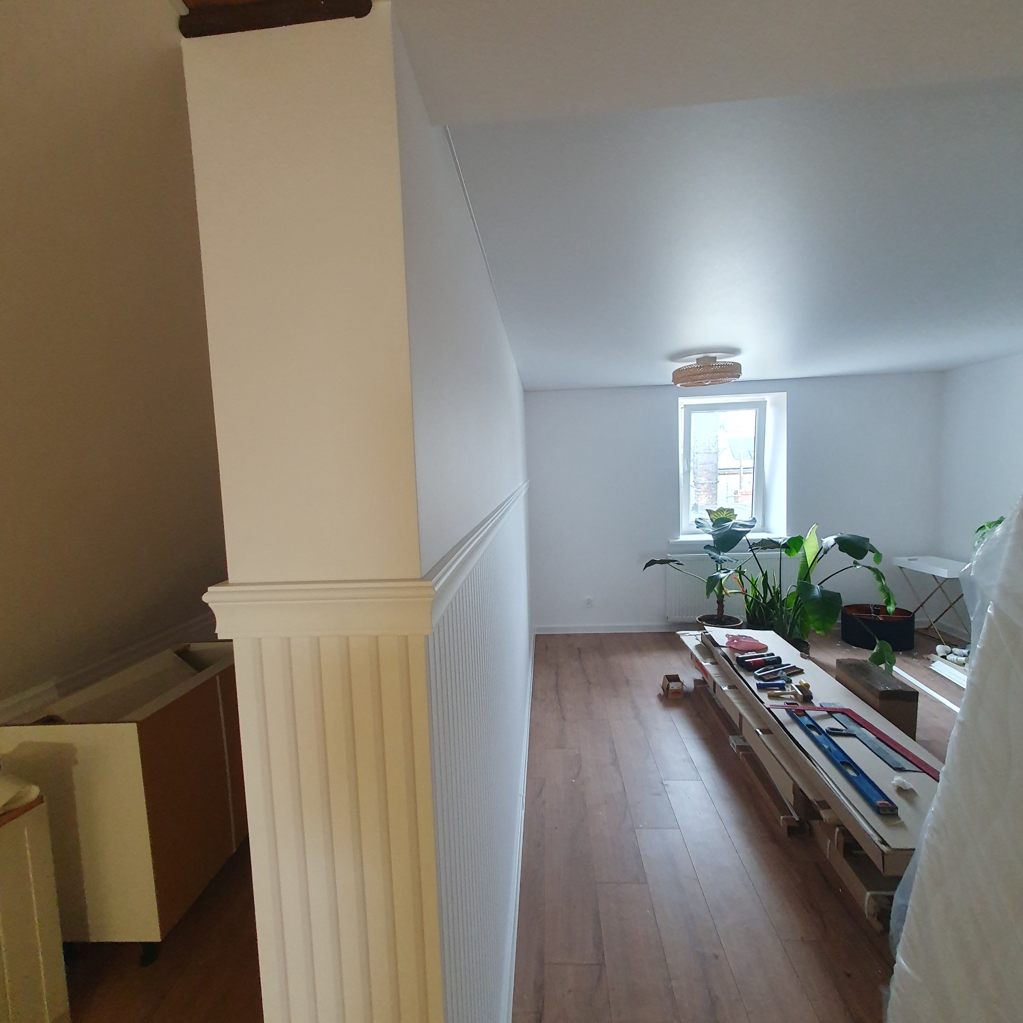 Białe ściany z dekoracyjną boazerią w trakcie malowania, widoczne narzędzia i materiały malarskie na podłodze, przygotowania do wykończenia wnętrza.