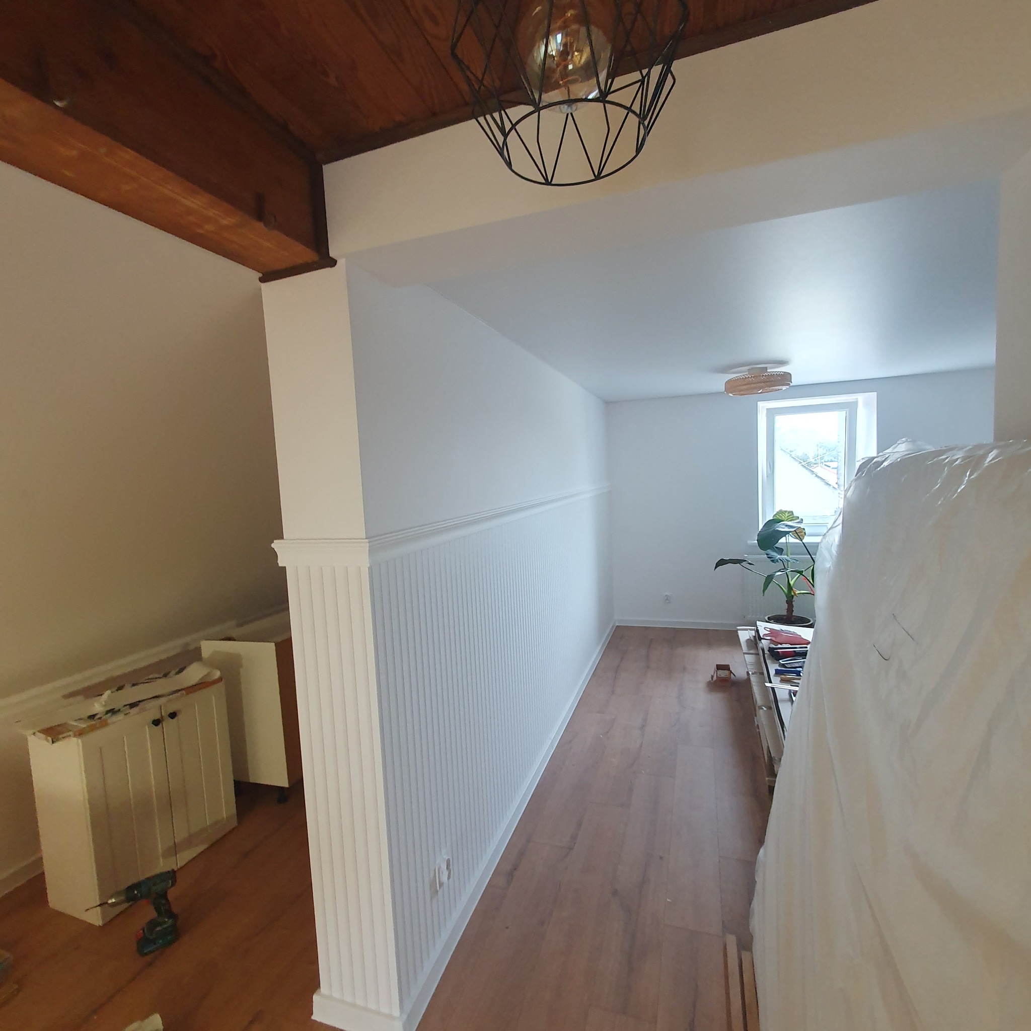 Wnętrze po remoncie: białe ściany z dekoracyjną listwą przypodłogową i pionowymi panelami, drewniana podłoga, widoczna wiertarka przy szafce, lampa sufitowa z geometrycznym kloszem i roślina...