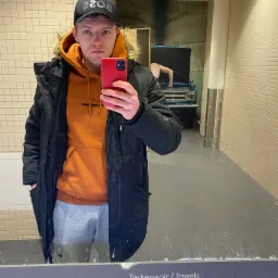 Mężczyzna w czapce i pomarańczowej bluzie robi selfie w łazience z kafelkami, w tle widoczny plakat z samochodem.