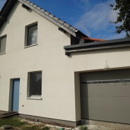 Remont Karol Cieśla - Jasny dom z garażem po renowacji elewacji. Widoczne okna z roletami, błękitne drzwi wejściowe i szara brama garażowa. Elewacja w odcieniach bieli, dach ciemny.