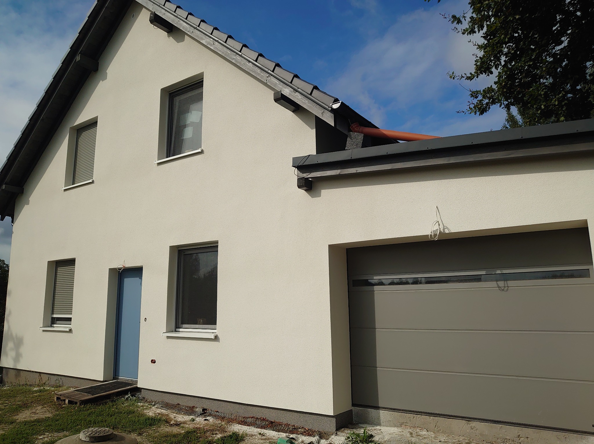 Jasny dom z garażem po renowacji elewacji. Widoczne okna z roletami, błękitne drzwi wejściowe i szara brama garażowa. Elewacja w odcieniach bieli, dach ciemny.
