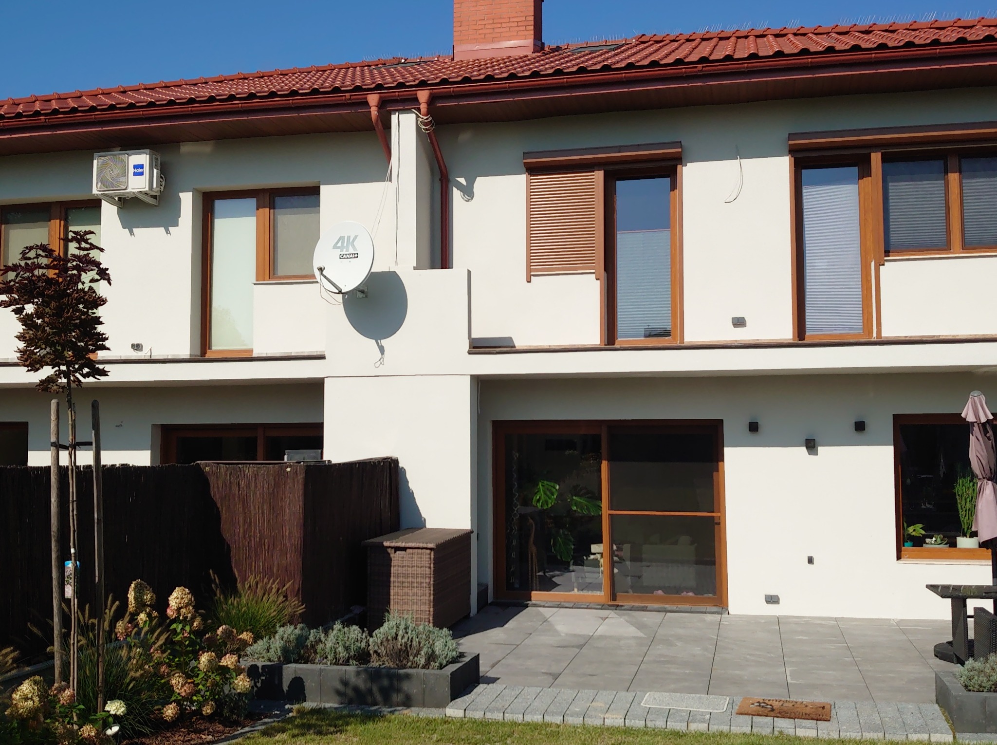 Jasna elewacja domu jednorodzinnego z brązowymi oknami i roletami, widoczny klimatyzator, antena satelitarna i zadbany ogród. Ujęcie w słoneczny dzień.