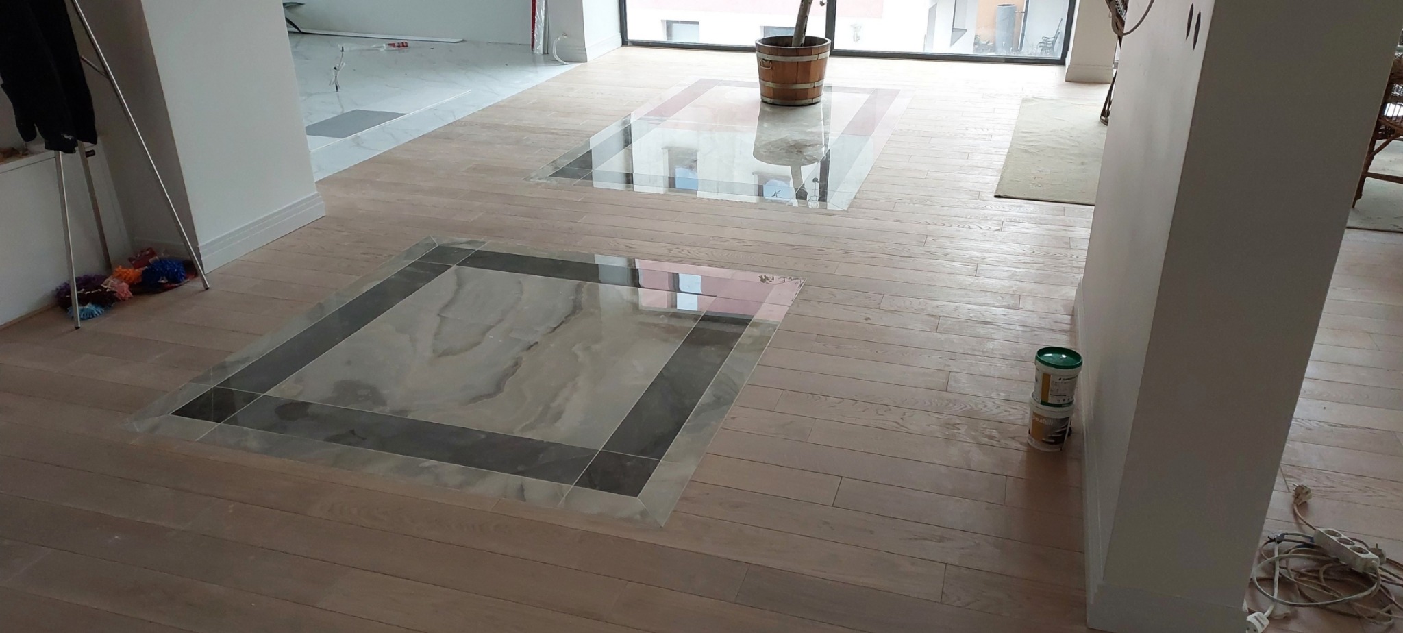 Jasna drewniana podłoga z wkomponowanymi kwadratami z marmuru, widoczne puszki z farbą przy ścianie i wiadro z rośliną w tle, wnętrze w trakcie wykańczania.